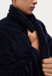 Granatowy polarowy szlafrok z wysokim kołnierzem i teksturowanym materiałem. Posiada dopasowane rękawy i rękę trzymającą kołnierz.