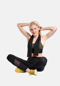 Black sports scarf worn over an olive green sports bra and black leggings with mesh accents. Yellow athletic shoes complete the outfit.
