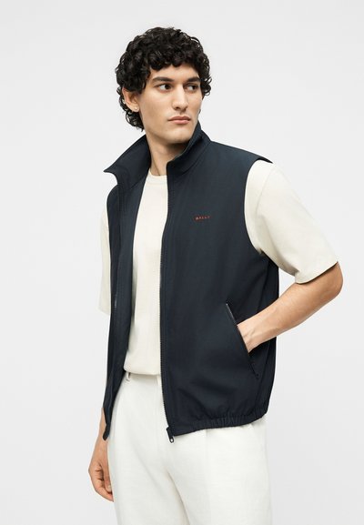 Άνδρας που φοράει μαύρο αμάνικο γιλέκο με φερμουάρ πάνω από μια κρεμ κοντομάνικη μπλούζα, με το ένα χέρι στην τσέπη του γιλέκου, κοιτάζοντας προς τα πλάγια.