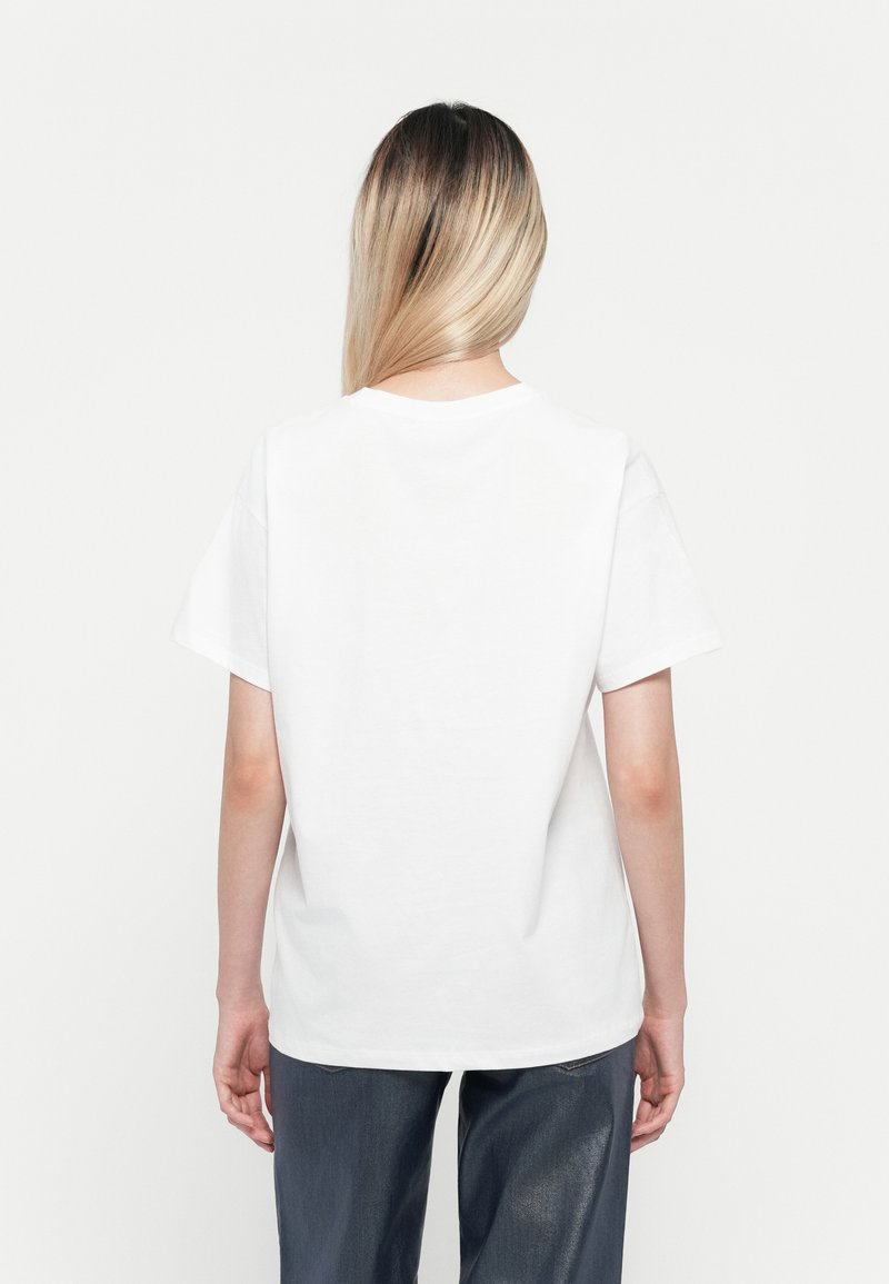 Persona con lunghi capelli biondi lisci, che indossa una maglietta bianca a maniche corte e pantaloni scuri, in piedi di spalle su uno sfondo bianco.