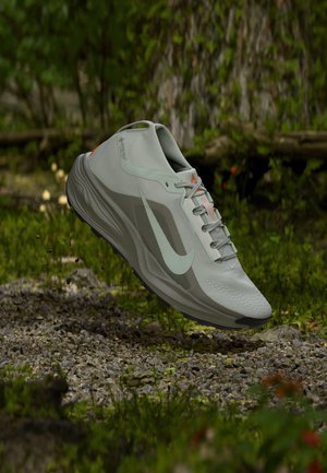 Pelēks Nike skriešanas kurpes ar baltu swoosh, lidojot pāri grants un sūnainai zemeslodei meža ainavā.