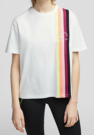 Biały t-shirt z krótkim rękawem z pionowymi tęczowymi paskami po prawej stronie i małym nadrukowanym tekstem, noszony z czarnymi legginsami.