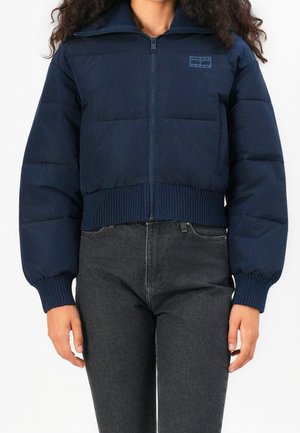 Frau trägt eine marineblaue Steppjacke mit gerippten Bündchen und Saum, die hochgeschlossen ist, kombiniert mit hochtaillierten schwarzen Jeans, stehend vor einem weißen Hintergrund.