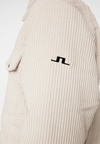 Beige kordjacka med ribbad textur, med en svart logotyp på axeln, och en lockficka på vänster sida.