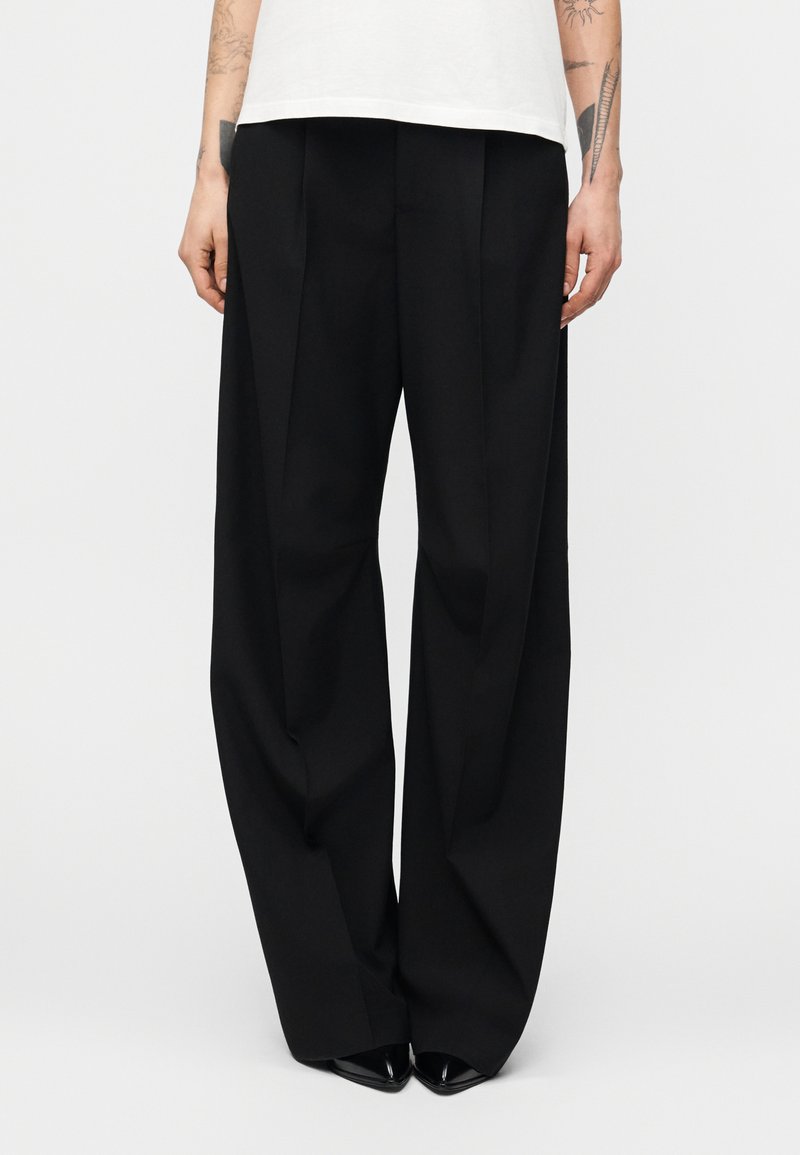 Άτομο που φοράει μαύρο παντελόνι μ ε φαρδιά μπατζάκια και μαύρα παπούτσια με μυτερή μύτη, στέκεται μπροστά σε ένα απλό λευκό φόντο.