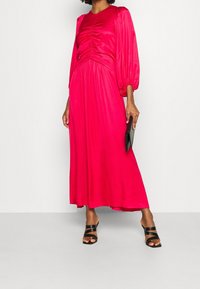 Kvinna i en lång, ljusrosa satinklänning med drapering i livet, puffärmar, svarta remsandaler och håller en svart clutchväska.