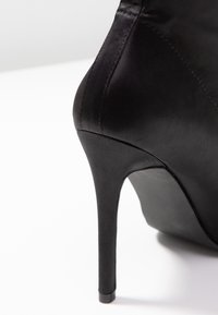 Chaussure à talon haut noire avec une finition satinée lisse, un bout pointu et un talon aiguille fin. Les coutures sont soigneusement réalisées.