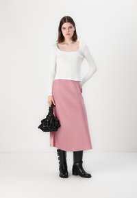 T-shirt blanc à manches longues côtelées, jupe maxi rose et bottes noires mi-mollet. Sac noir texturé avec un design froncé. Fond blanc uni.