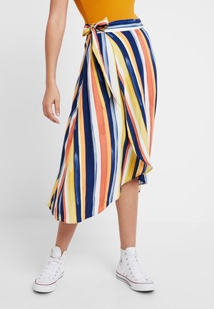 Kvinde iført et flerfarvet stribet omslagsskjørt bundet i taljen, kombineret med hvide høje sneakers og en sennepsfarvet top.