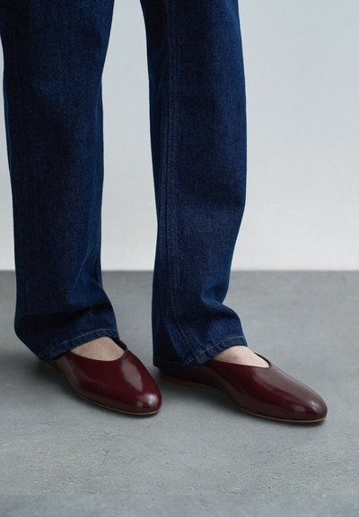 Sapatos de couro borgonha sem atacadores, com biqueira arredondada e design recortado, usados com jeans denim azul, apresentando um corte reto nas pernas.