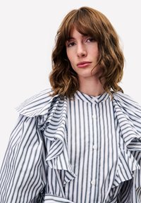 Junge Frau mit welligem braunem Haar, die eine weiße Bluse mit schwarzen vertikalen Streifen und Rüschendetails an den Schultern trägt, vor neutralem Hintergrund.