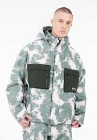 Kapuzensh jacket mit einem grünen und weißen Wolkenmuster, zwei mit Reißverschluss versehenen Brusttaschen und einem lockeren Schnitt. Das Material scheint wetterbeständig zu sein.