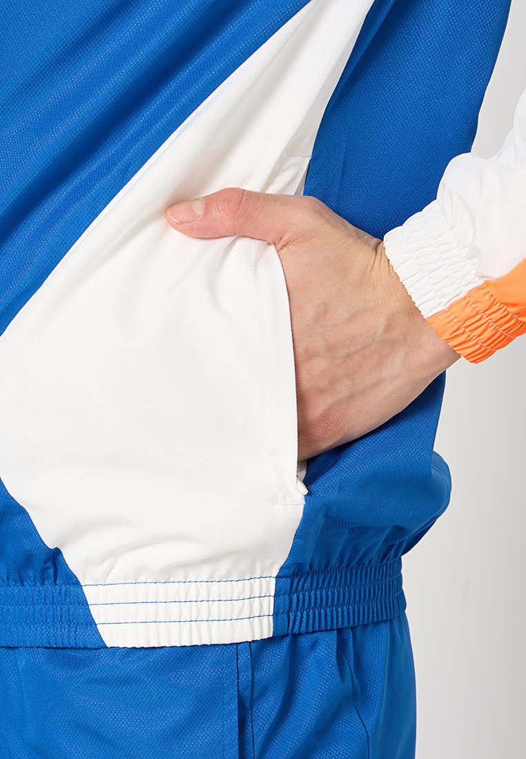 Chaqueta azul y blanca con un tejido texturizado, que presenta un bolsillo lateral y puños elásticos. Contraste de color notable con detalles en naranja.