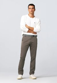 Maglione bianco a trecce, pantaloni grigi e una cintura con motivo. Abbinato a scarpe sneakers chiare. Lo sfondo neutro mette in evidenza i dettagli dell'outfit.