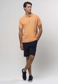 heleoranž värvi polo-särk kraega ja lühikeste varrukatega, sobitatud tumesiniste lühikeste pükstega. Komplekti täiendavad tumedad tossud.