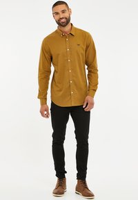 Braunes Hemd mit Knöpfen aus einem weichen Stoff, mit einem Button-Down-Kragen und einer Vordertasche, kombiniert mit schwarzen Slim-Fit-Jeans und braunen Stiefeln.