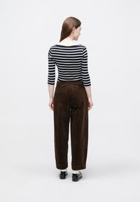 Žena otočená chrbtom, oblečená v čiernobielom pruhovanom tričku, hnedých širokých manžestrákových nohaviciach, bielych ponožkách a čiernych topánkach na bielom pozadí.