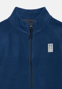 Tumed sinine fliisjakki kõrge kraega, esime must tõmblukk ja väike hall logopatch, millel on ümmargused detailid vasakul küljel.