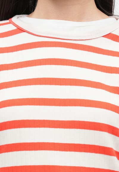 Κοντινό πλάνο ενός ατόμου που φοράει ένα ριγέ λευκό πουκάμισο με οριζόντιες πορτοκαλί ρίγες και λευκή στρογγυλή λαιμόκοψη.