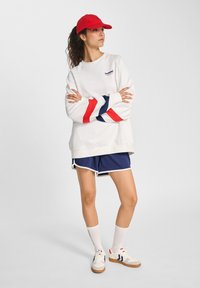 Vrouw in een witte sweatshirt met rode en marineblauwe strepen, marineblauwe shorts, witte sokken, sneakers en een rode pet, staand met de armen gekruist.