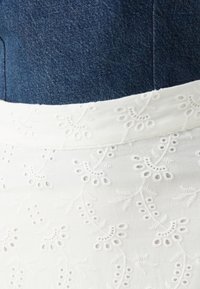 Falda blanca bordada con patrones florales y detalles de ojal; combinada con una blusa de mezclilla azul. Tejido suave con una textura lisa.