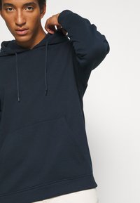 Marineblaue Kapuzenpullover aus weichem Material mit einer Vorderentasche und Kordelzugkapuze. Mit langen Ärmeln und gerippten Manschetten.