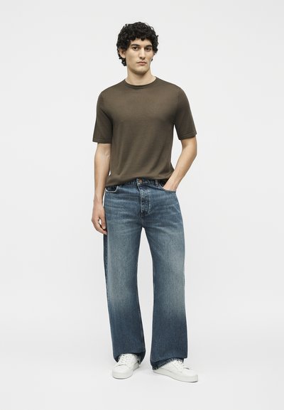 Νέος άνδρας με κατσαρά μαλλιά, φορώντας ένα ελιά κοντομάνικο μπλουζάκι, φαρδιά ξεβαμμένα μπλε τζιν και λευκά αθλητικά παπούτσια, στέκεται με το ένα χέρι στην τσέπη.