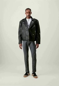 Svart läderbikerskinnjacka med dragkedjor och fickor, buren över en vit t-shirt, kombinerad med grå byxor och svarta sneakers på en neutral bakgrund.