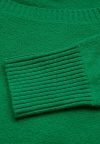 Grön tröja med mjukt, texturerat tyg och ribbade manschetter. Den övergripande designen är enkel, med en slät yta genom hela plagget.