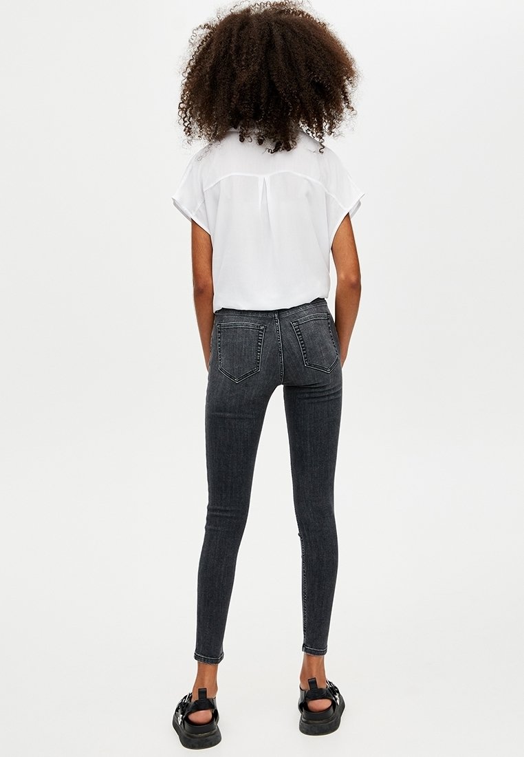 Jeans skinny negros de cintura alta, con cinco bolsillos y un diseño limpio, combinados con una blusa blanca de corte holgado y sandalias negras.