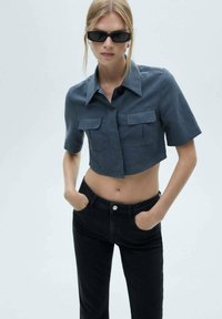 Abgeschnittenes dunkelblaues Hemd mit Kragen, kurzen Ärmeln und zwei vorderen Taschen, kombiniert mit hoch taillierten schwarzen Jeans. Glatte Stofftextur.