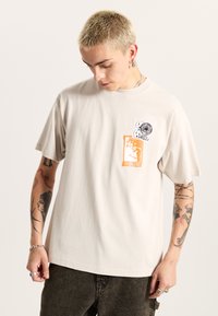 Helle beigefarbene Baumwoll-T-Shirt mit kurzen Ärmeln, das im Brustbereich über drei grafische Aufnäher in Schwarz, Orange und Weiß verfügt.