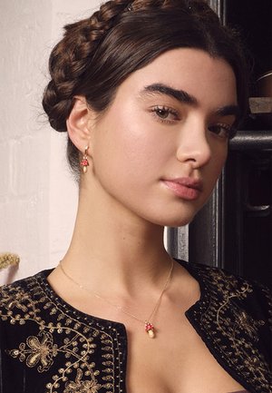Young woman with braided hair wearing black embroidered jacket, gold mushroom pendant necklace, and matching mushroom-shaped earrings.