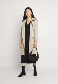 Beige trenchcoat over en svart ribbestrikket kjole, kombinert med svarte ankelstøvler og en teksturert svart håndveske. Rent, moderne design.