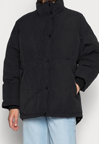 Persona vestida con una chaqueta puffer negra abotonada y de cuello alto, con las manos parcialmente visibles, combinada con unos vaqueros de mezclilla azul claros.