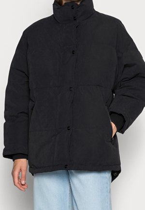 Una persona che indossa un parcobuto nero con bottoni e colletto alto, con le mani parzialmente visibili, abbinato a jeans di denim azzurri chiari.