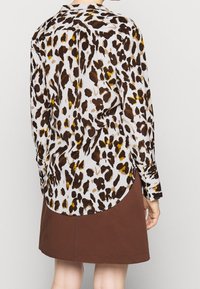 Vit långärmad skjorta med knappar, prydd med brun och gul leopardtryck, med en rundad nederkant och uppvikta ärmar. Bärs tillsammans med en brun kjol.