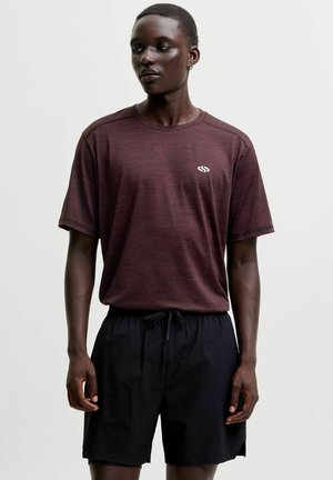 Homme portant une chemise bordeaux à manches courtes et un short noir avec cordon, debout devant un fond clair uni.