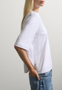 Una persona bionda con una camicia bianca a maniche corte e jeans blu, con una mano in tasca, rivolta di lato su uno sfondo neutro.