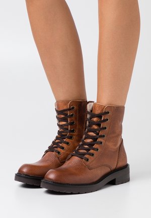 Botines marrón de cuero con cordones y suelas negras, usados en piernas desnudas sobre un fondo liso y claro.