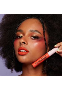 Läppprodukt med en glansig orange nyans i en matt, cylindrisk tub med ett rosa lock och fet svart text. Huden har en strålande finish.