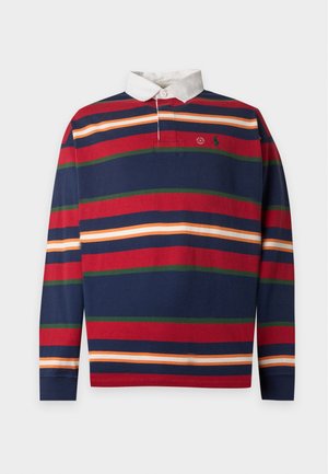 Jeune homme portant une chemise à col à rayures bleu marine, rouge et orange à manches longues, un pantalon cargo beige et des baskets blanches, debout avec une main dans la poche.