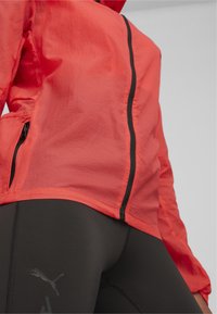 Rote leichte Jacke mit strukturiertem Finish, ausgestattet mit einem durchgehenden Reißverschluss und schwarzen Akzenten, kombiniert mit figurbetonten schwarzen Leggings mit einem Logo.