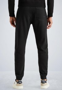 Mies, joka käyttää mustia jogger-housuja, joissa on takatasku, ja valkoisia tossuja beige-akkenteilla, seisoo harmaalla lattialla.