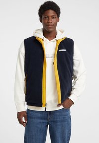 Marineblaues Fleeceweste mit gelben Reißverschlüssen und Akzenten, versehen mit einem Logo-Patch auf der Brust, kombiniert mit einem weißen Kapuzenpullover und blauen Jeans.