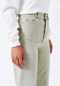 Jeans Verde Chiaro a Vita Alta con Design a Gamba Dritta e Finiture in Ottone Rame. Mostrano Denim Testurizzato e Tasche Laterali.
