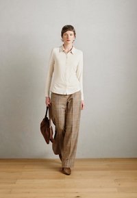 Helles beiges Hemd mit Knöpfen, kombiniert mit weit geschnittenen karierter Hose. Das Model trägt eine braune Handtasche und braune Schuhe. Einfarbiger Hintergrund.