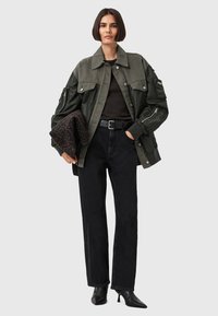 Giacca verde oliva con design oversize e tasche applicate, abbinata a una maglia nera, jeans neri a vita alta e tacchi a punta neri.