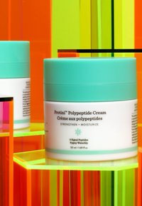 White jar of Protini Polypeptide Cream with a turquoise lid, sitting on colorful acrylic blocks. Text details visible on the jar.