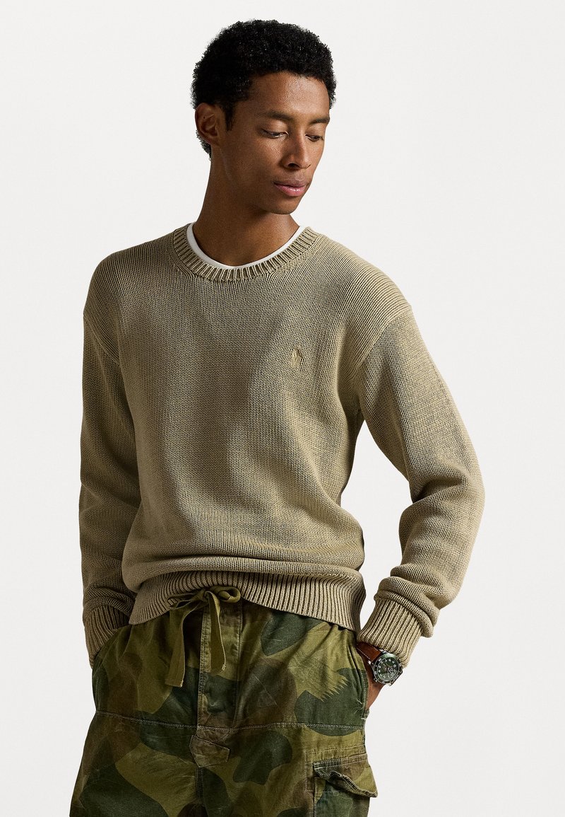 Beżowy sweter dzianinowy z prążkowanym dekoltem i mankietami, noszony na białej koszulce. W komplecie z zielonymi spodniami cargo w kamuflażu i zegarkiem.